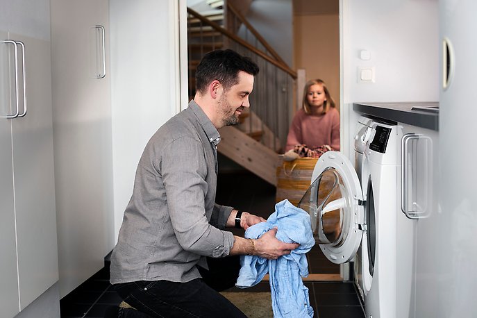 En man står på huk framför en öppen tvättmaskin och stoppar in tvätt i maskinen. I bakgrunden syns en flicka som är på väg in till mannen.