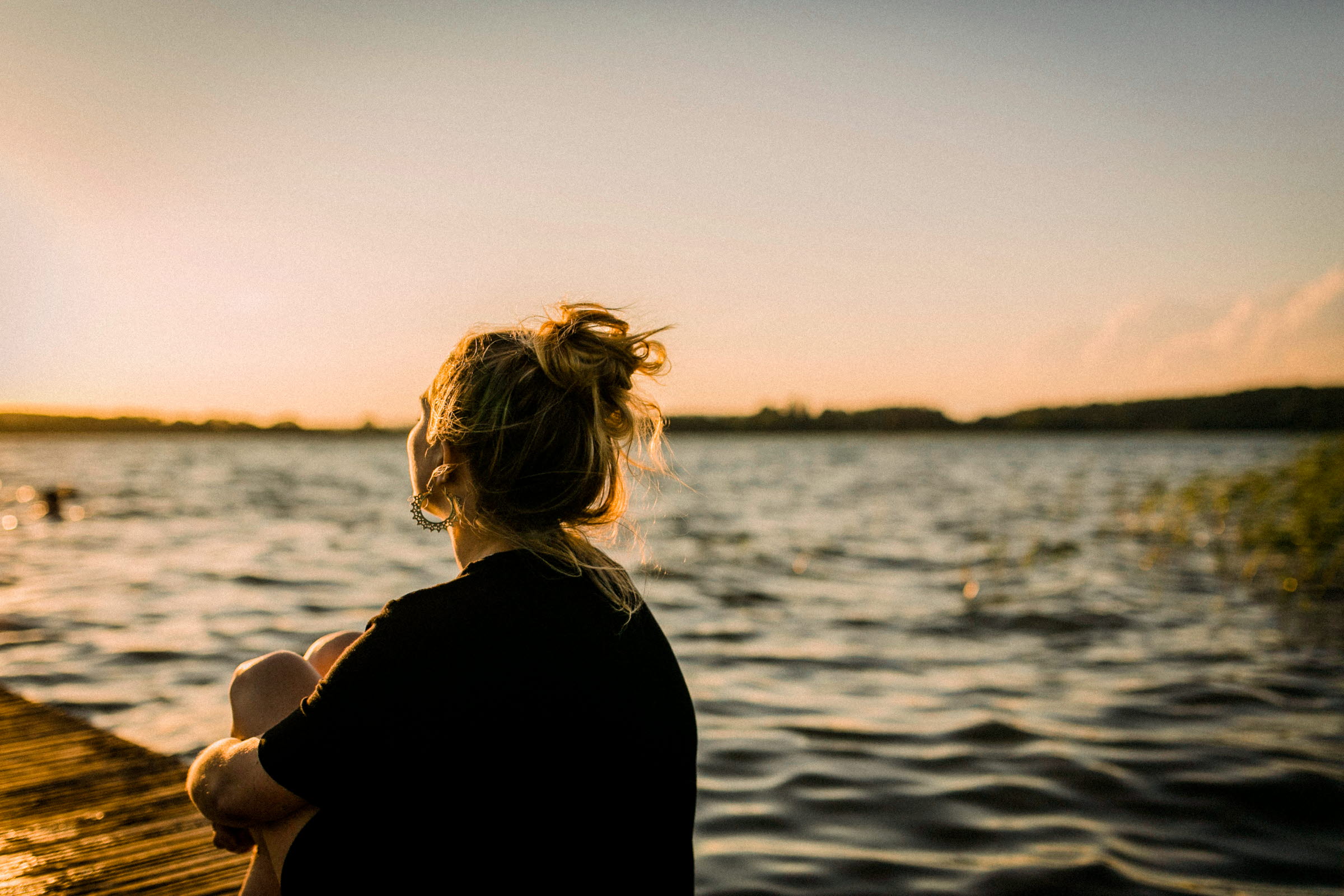En kvinna i svart tröja och uppsatt hår sitter på en brygga och blickar ut över en sjö.