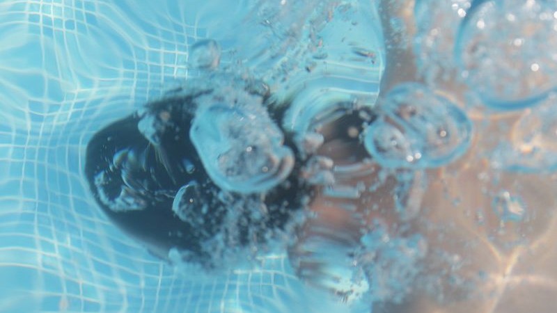 Närbild rakt ner i en pool där man ser bakhuvudet på en människa som simmar under vattnet. Ovanför huvudet kommer luftbubblor upp.