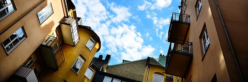 Foto taget upp mot en blå himmel med vita moln. Runtomkring finns höga husväggar med fönster och balkonger. 
