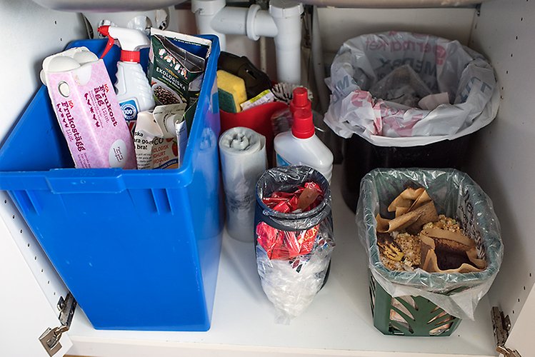 Skåpet under en diskho. En stor plastlåda till vänster innehåller plats och pappersförpackningar. Till höger finns en soptunna samt ett kärl med matavfall. I mitten står en liten behållare med mjuka plastförpackningar, en rulle matavfallspåsar samt städmaterial.